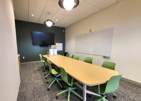 Photo of conference room with table, chairs, whiteboard, and monitor visible.