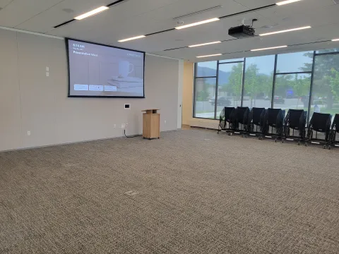 Large room with projector, screen, podium, and chairs stacked on carts by the window on the right hand side. 