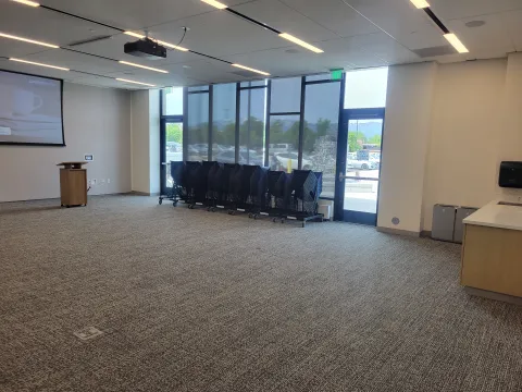 Large room with projector, screen, podium, and chairs stacked on carts by the window on the right hand side. 